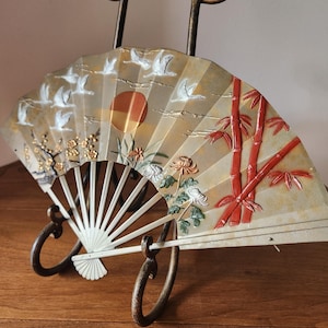 May include: An ornate, open fan with a cream-colored background, featuring a painted scene of flying birds, a setting sun, and bamboo stalks. The fan is displayed on a decorative metal stand, showcasing its intricate design and artistry.