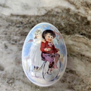 May include: A white ceramic Easter egg with a painted scene of two children. One child is riding a bicycle, and the other is standing behind them. The egg has a glossy finish and is decorated with pastel colors.