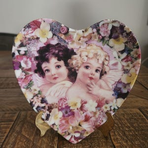 May include: Heart-shaped decorative plate featuring two cherubs surrounded by a floral design in shades of pink, purple, yellow, and white. The plate is displayed on a wooden stand, showcasing a vintage aesthetic.