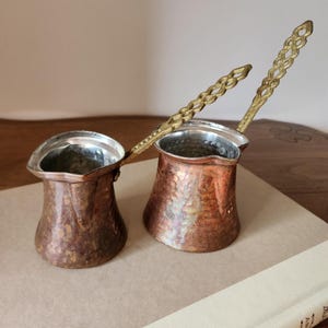 May include: Two copper Turkish coffee pots with brass handles. The pots have a hammered texture and a silver interior. The handles are ornate and extend from the pots. The pots are sitting on a light brown surface.