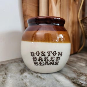 May include: A round ceramic bean pot with a glossy finish, featuring a dark brown rim and a cream-colored base. The text BOSTON BAKED BEANS is printed in dark brown, stylized lettering on the front of the pot, which has a wide body and a narrow opening.