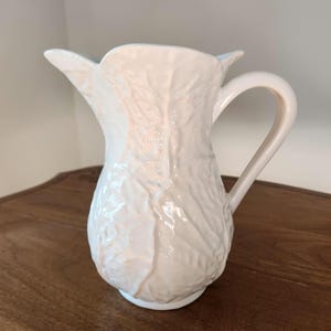 May include: A cream-colored ceramic pitcher with a textured, leaf-like design. The pitcher has a curved handle and a spout. The pitcher is sitting on a wooden surface.