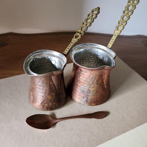 May include: Two copper Turkish coffee pots with ornate gold-colored handles and a matching spoon. The pots have a hammered texture and silver-colored interiors. The spoon is a similar copper color. The items are on a neutral-colored surface.