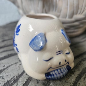 May include: A small, cream-colored ceramic pig-shaped planter or container featuring a glossy finish with blue hand-painted floral accents on the sides, blue ears, and a blue-and-white checkered pattern on the snout area, with a smooth, rounded body.
