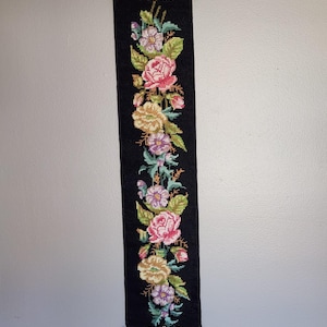 May include: A black, vertical tapestry with a floral cross-stitch design. The embroidery features pink, yellow, and purple flowers with green leaves. The tapestry is framed with ornate, gold-colored metal hardware at the top and bottom.