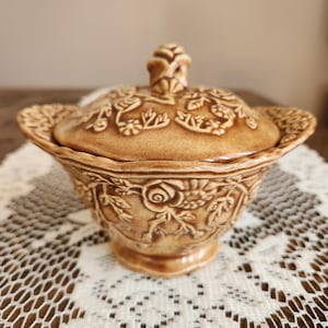 May include: A tan ceramic lidded serving dish with embossed floral and leaf designs. The dish has a pedestal base and handles. The lid features a decorative knob. The dish is sitting on a white lace tablecloth.