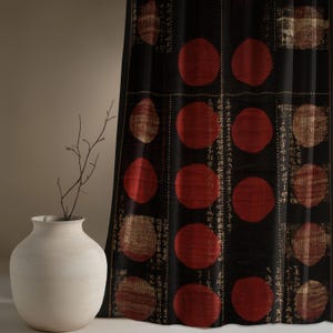 May include: A white ceramic vase holds a slender, bare branch. A black curtain with large red circles and faded beige squares is in the background. The curtain has vertical columns of small, faded text.
