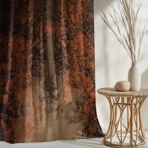 May include: A patterned curtain with a blend of brown, orange, and tan hues. The design features a faded, ornate pattern. A small, round, woven side table holds a white vase, a small tan pot, and dried grass.