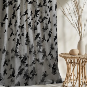 May include: A light gray curtain with a repeating pattern of black birds in flight. The birds are depicted with their wings spread, creating a dynamic and airy feel. The curtain is next to a small, round, woven side table with a vase of dried plants.