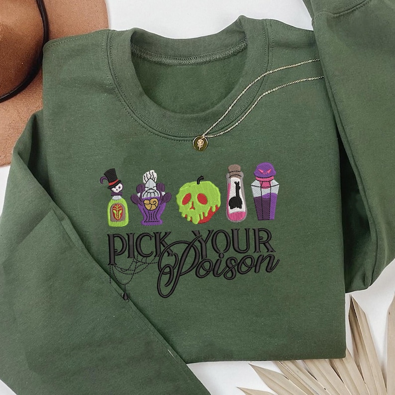 May include: A green sweatshirt with the phrase "Pick Your Poison" and embroidered images of potion bottles and a skull-shaped apple. A gold necklace with a small pendant is draped on the sweatshirt.