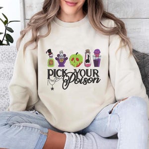 May include: A cream-colored sweatshirt featuring the phrase "Pick Your Poison" in black script, with a spider web and spider graphic. The design includes colorful potion bottles and a green apple with a skull face.