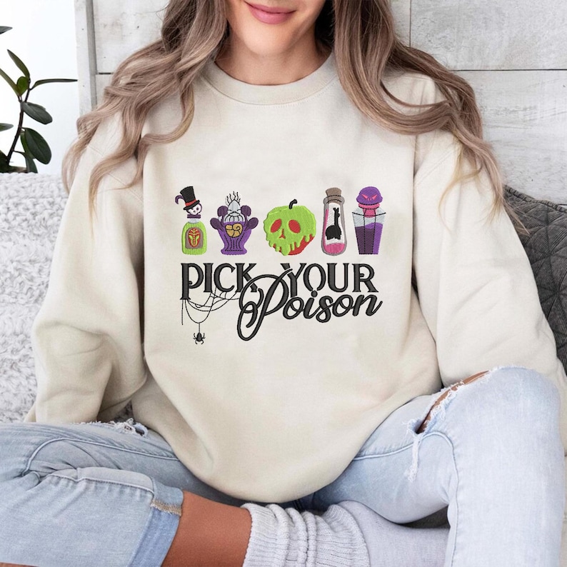May include: A cream-colored sweatshirt featuring the phrase "Pick Your Poison" in black script, with a spider web and spider graphic. The design includes colorful potion bottles and a green apple with a skull face.