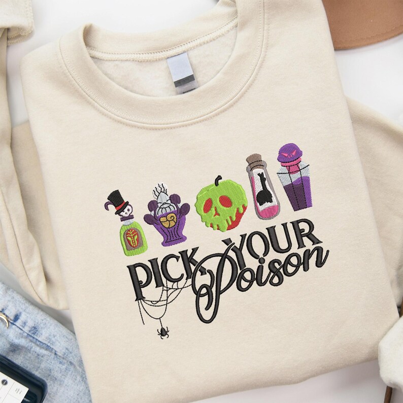 May include: Cream-colored sweatshirt with an embroidered design featuring potion bottles and a green apple with a skull face. The text "PICK YOUR POISON" is written in black script with a spider web and spider detail.