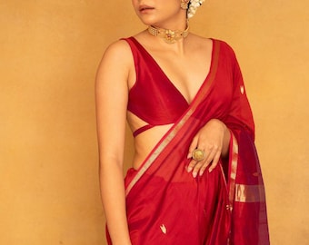 May include: A woman wearing a red sari with gold trim and a matching red blouse. She is adorned with gold jewelry, including a choker necklace and a large ring. Her hair is styled up with white flowers.