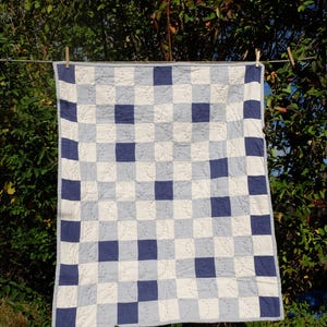 Peut inclure: Une courtepointe faite à la main avec un motif en damier de carrés blancs, bleu clair et bleu marine. La courtepointe est suspendue à une corde à linge à l'extérieur, avec un fond de feuillage vert et un ciel bleu clair. La courtepointe mesure environ 100 cm sur 100 cm.