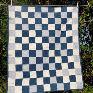 Könnte beinhalten: Eine handgefertigte Steppdecke mit einem Schachbrettmuster aus marineblauen, hellblauen und weißen Quadraten. Die Steppdecke hängt an einer Wäscheleine mit Holzklammern, die ihr Design und ihre Handwerkskunst zeigen. Die Steppdecke hat eine quadratische Form.