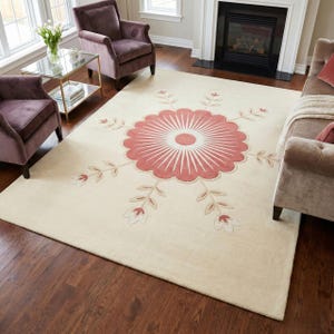 Peut inclure: Un tapis de sol crème avec un grand motif floral stylisé dans des tons de rose et de beige. Le tapis présente une fleur centrale avec des lignes rayonnantes et des accents floraux sur les bords. Le tapis est posé sur un parquet foncé.