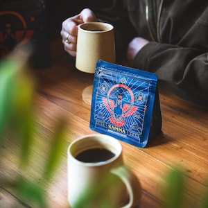 Może przedstawiać: Niebieska torebka na kawę z czerwono-białą grafiką ptaka i szczytów górskich, z napisem Kamma Himalayan Coffee. Torebka posiada zapięcie strunowe. Obok znajdują się dwa beżowe ceramiczne kubki wypełnione ciemną kawą.