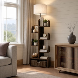 May include: A dark brown floor lamp with a built-in bookshelf. The lamp has a beige lampshade and a series of shelves shaped like curved brackets. The shelves hold books, plants, a clock, and decorative items. A woven basket is at the base.