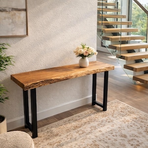 Puede incluir: Una consola de madera con una tapa de borde vivo y patas de metal negro. La mesa mide aproximadamente 152 cm de largo y está coronada por un jarrón blanco con flores. La mesa está colocada contra una pared blanca.