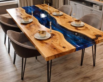 May include: A wooden dining table with a blue resin river design. The table has a natural wood grain finish and is supported by black metal hairpin legs. The table is set with plates, bowls, and glasses, and surrounded by gray chairs.