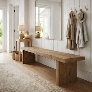 May include: A rustic wooden bench with a natural finish sits in a hallway. The bench is rectangular and has thick legs. A beige rug, a side table with lamps, and a mirror are also visible. Coats and a hat hang on the wall.