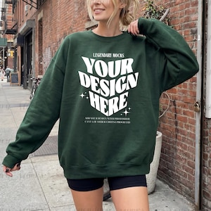 May include: A dark green sweatshirt with the text "YOUR DESIGN HERE" in white, with the words "LEGENDARY MOCKS" above and additional text below. The sweatshirt is worn by a person standing on a city sidewalk.