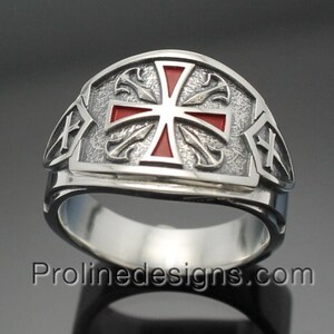 Könnte beinhalten: Ein silberner Ring mit einem roten Kreuzdesign. Das Kreuz ist von einem dekorativen Rand umgeben. Der Ring ist ein Statement-Stück und wäre ein tolles Geschenk für jemanden, der religiös ist.