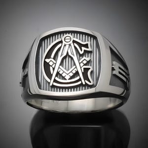 May include: A silver Masonic ring featuring a black and white square and compass design. The ring has a textured band.