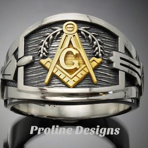 May include: A silver ring with a gold Masonic symbol, featuring a compass and square, a 'G' in the center, and a wreath of leaves around the symbol.