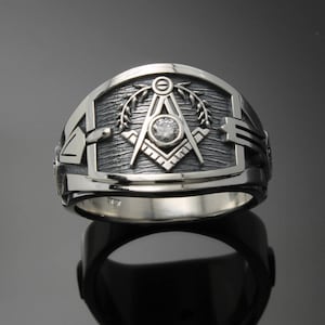 May include: A silver Masonic ring with a square and compass design and a clear stone in the center. The ring has a textured band and a detailed design.