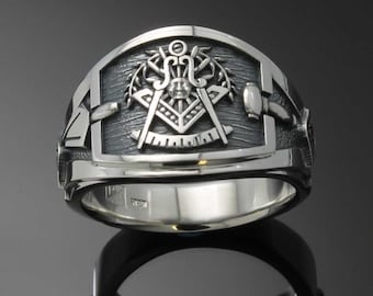 May include: A silver Masonic ring with a detailed design featuring a compass, square, and other symbols. The ring has a textured band and a raised design.
