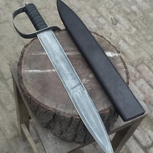 May include: A steel sword with a double-edged blade and a black ribbed handle featuring a metal hand guard. The sword is accompanied by a matching black scabbard. The blade has a polished finish with visible metal grain, tapering to a sharp point.
