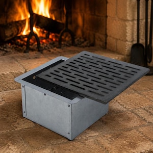 May include: A rectangular metal fireplace humidifier box with a matte grey finish and exposed screw fasteners. The unit features a removable black cast iron grate lid with a series of horizontal ventilation slots for steam release.