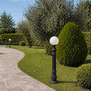 Può includere: Un lampione nero da esterno con una luce a globo bianca. Il lampione è ornato con dettagli decorativi e si trova su un prato verde accanto a un sentiero di pietra. Lussureggianti alberi e cespugli verdi sullo sfondo.
