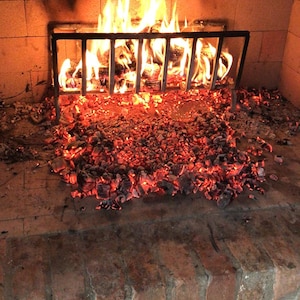 Puede incluir: Una chimenea con un fuego ardiente. Las llamas se elevan de los troncos en llamas en una rejilla de metal negro, rodeadas de brasas brillantes. La mampostería de la chimenea es visible, con una pila de leña en la parte inferior.