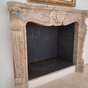 Könnte beinhalten: Ein beiger Marmorkamin mit aufwendigen Schnitzereien und dunklem Innenraum. Der Kamin hat einen dekorativen Mantel mit floralen und scrollartigen Details. Der Feuerraum hat einen Metallrost am Boden. Der Kamin steht vor einer weißen Wand.