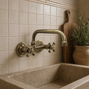 May include: Antique brass wall-mounted faucet with cross handles. The faucet has a curved spout and is mounted on a tiled wall. A wooden cutting board and a potted plant are visible in the background.