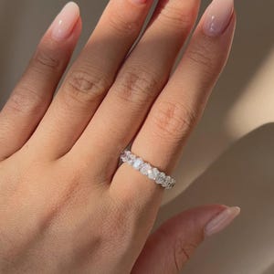 Puede incluir: Una mano con uñas largas y pulidas que lleva un anillo de plata. El anillo presenta una banda continua de piedras preciosas claras de talla ovalada. Las piedras preciosas están muy juntas, creando una apariencia brillante y elegante. Las uñas son de un rosa suave y natural.