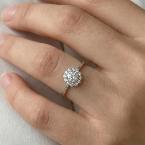May include: A silver ring with a round, sparkling center stone surrounded by smaller stones. The ring is set on a thin band and is shown on a person's finger.