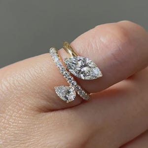 May include: Three gold rings with diamond accents. The largest ring features a marquise-cut diamond. A second ring has a pear-shaped diamond. The third ring is a band with small diamonds. The rings are set on a finger.