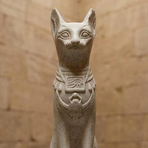 Puede incluir: Una estatua de piedra moteada de color claro que representa un gato sentado de estilo egipcio. El gato lleva un collar con un colgante rectangular con el Ojo de Horus y un emblema de escarabajo alado centrado en el pecho, con orejas puntiagudas.
