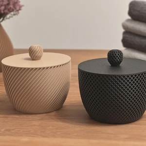 Pode incluir: Dois recipientes decorativos com tampa. Um é bege com um design em espiral, o outro é preto com um padrão de losangos. Ambos têm um botão esférico no topo. Os recipientes estão numa superfície de madeira.