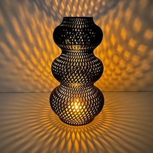 Peut inclure: Une lanterne en métal noir avec un design empilé unique. La lanterne présente un motif en treillis qui projette des motifs complexes d'ombre et de lumière. La lueur chaleureuse de la lumière crée une ambiance chaleureuse. Un élément décoratif.
