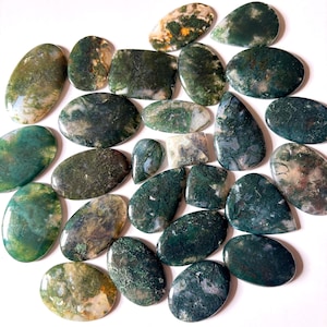 Puede incluir: Una colección de piedras preciosas de ágata musgosa pulidas en varias formas, incluyendo ovaladas, de lágrima y rectangulares. Las piedras muestran una mezcla de colores verdes, blancos y marrones, con patrones intrincados que se asemejan al musgo. Las piedras están dispuestas sobre una superficie blanca.