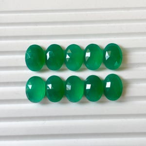 Puede incluir: Diez gemas verdes ovaladas y facetadas dispuestas en dos filas de cinco. Las gemas tienen una superficie pulida y un color verde vibrante. Las piedras están sobre un fondo blanco con estrías.