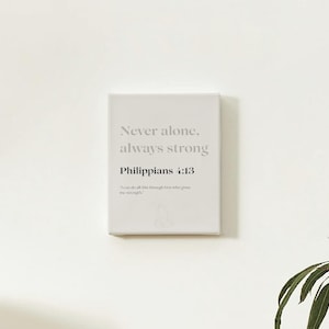 Könnte beinhalten: Rechteckiger Leinwanddruck mit dem Text "Never alone, always strong" und "Philippians 4:13" in Grau. Das Zitat "I can do all things through him who gives me strength" ist ebenfalls enthalten. Der Druck ist an einer weißen Wand befestigt.