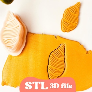 May include: An overhead shot shows a piece of orange clay with a seashell-shaped imprint and two separate seashell cutouts. A 3D printing file is available. The clay is on a white surface.