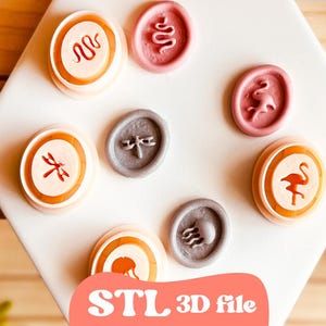 Könnte beinhalten: Eine Sammlung von Wachssiegeln in verschiedenen Farben und Designs, darunter eine Schlange, eine Libelle und ein Flamingo. Die Siegel sind auf einer weißen Oberfläche angeordnet, mit dem Text "STL 3D file" in einem rosa Banner.