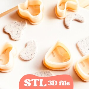 May include: A collection of light orange plastic cookie cutters in various shapes, including a heart, a crescent moon, and a square. The cutters are arranged on a white surface with speckled clay shapes. The text "STL 3D file" is visible.
